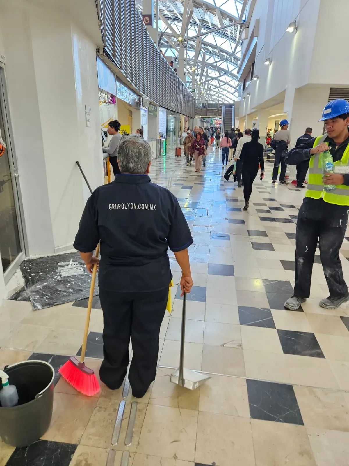Integrante de Grupo Lyon de espaldas con el uniforme GRUPOLYON.COM.MX barriendo un pasillo del aeropuerto